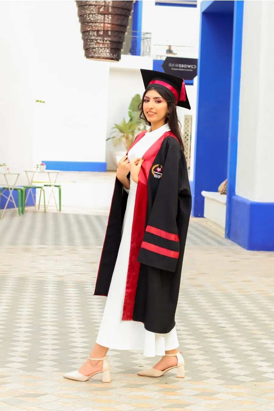 خرّيجة بروب التخرّج الأسود والأحمر واقفة في ساحة ببلاط هندسي وجدران زرقاء وبيضاء