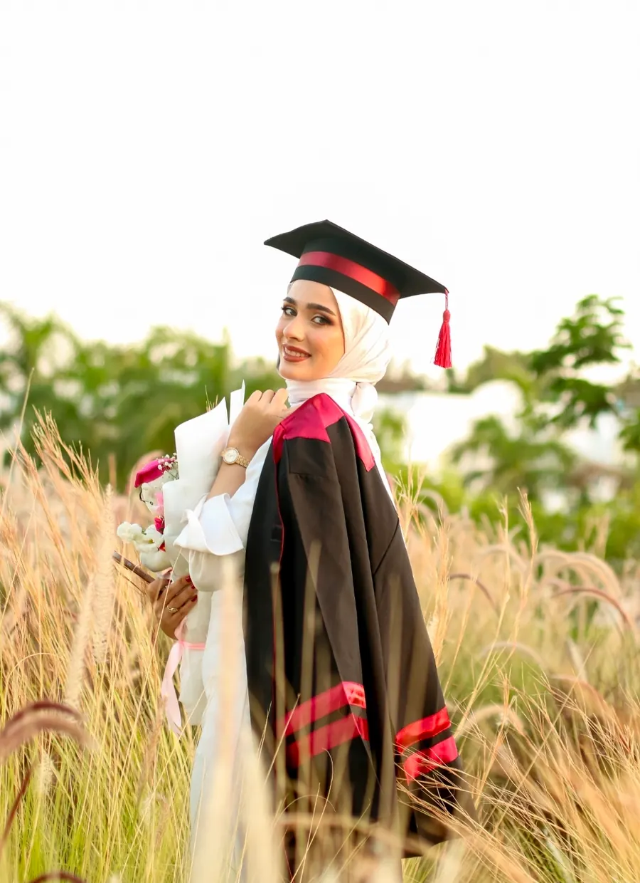 خرّيجة بروب التخرّج تحمل باقة ورد وتلتفت نحو الكاميرا وسط حقل أعشاب ذهبية عند الغروب