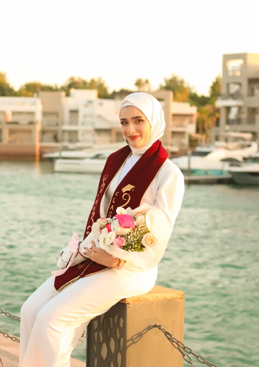 خرّيجة تحمل باقة ورد وتجلس أمام مرسى فيه قوارب ومياه فيروزية عند الغروب