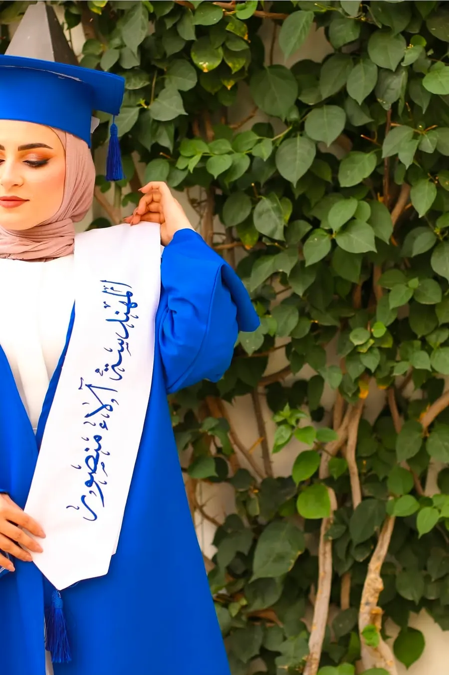 لقطة قريبة لشال تخرّج أبيض بخطّ عربي مع روب أزرق أمام أوراق شجر خضراء
